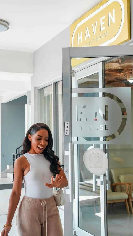 A woman happily standing at the entrance of Haven Beauty House, a modern beauty salon with glass doors, welcoming ambiance, and a cheerful atmosphere for clients.