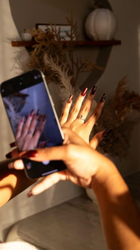 Gorgeous hand with manicured nails and ring, taking a selfie in a cozy, stylish space with decorative dried plants and modern decor. Perfect for beauty, manicure, and nail salon SEO.